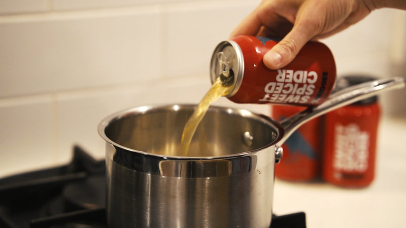 Person pouring cider from a can of mr bridges sweet spiced cider into a pot on a stove.